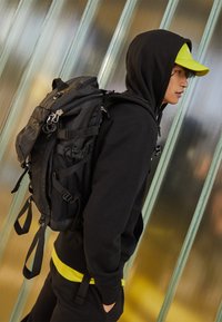 Czarny plecak z wieloma przegrodami, akcentami z siateczki i regulowanymi paskami. Nosi się go na czarnej stylizacji z żółtym detalem w pasie.