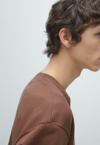 Bruine katoenen trui met ronde hals, heeft een gladde textuur en minimale stikdetails, getoond in een profiel van een model.