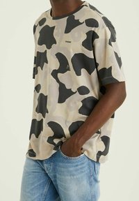 Camiseta de manga corta en beige con patrón de camuflaje negro y gris, cuello redondo, hecha de algodón suave y con un corte relajado.