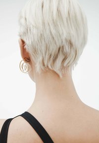 Pendientes de aro de oro y plata, con círculos entrelazados, que se llevan en un peinado corto y rubio claro sobre un fondo liso.