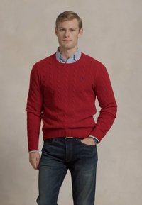 Raudonas megztinis su pynėmis, turintis apvalų kaklą, derinamas su šviesiai mėlynu marškinėlių švarkeliu, vilkimas su tamsiai mėlynomis džinsais. Išskirtinė logotipo detalė ant megztinio.