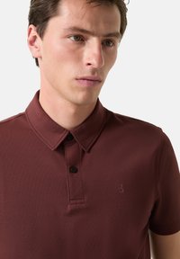 Bordová polo košile s klasickým límcem, dvěma černými knoflíky a malým logem na hrudi. Texturovaná tkanina s jemným vzorem.