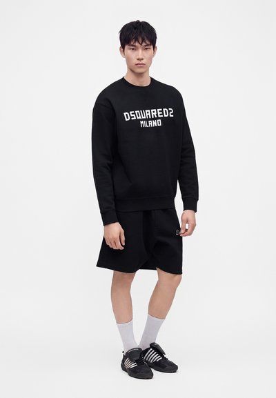 Νέος άνδρας με μαύρο φούτερ Dsquared2 Milano, μαύρα σορτς, λευκές κάλτσες και μαύρα αθλητικά παπούτσια στέκεται μπροστά σε απλό φόντο.