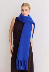 Vrouw in een mouwloze zwarte jurk en een oversized pluizige blauwe sjaal met franjes om haar hals, tegen een effen achtergrond.