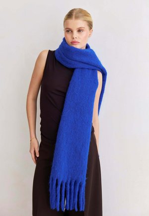 Femme portant une robe noire sans manches et une écharpe bleue moelleuse à franges enroulée autour du cou, sur fond uni.