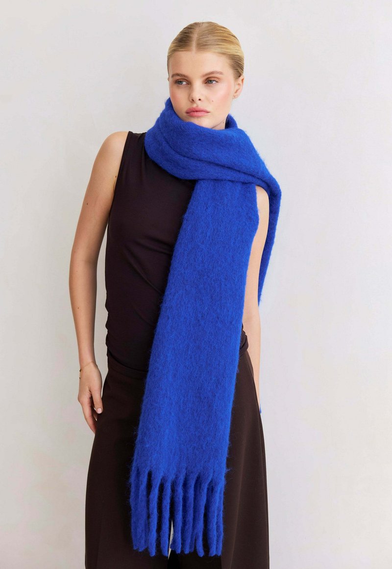Vrouw in een mouwloze zwarte jurk en een oversized pluizige blauwe sjaal met franjes om haar hals, tegen een effen achtergrond.
