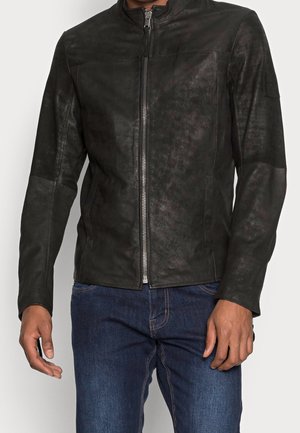 Homme portant une veste en cuir noire zippée avec un col montant et un jean bleu foncé, debout devant un fond clair uni.