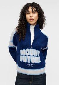 Μπλε πουλόβερ με φερμουάρ και έντονη επιγραφή "MOUNT FUTURE". Κατασκευασμένο από μαλακό πλεκτό υλικό με ανοιχτό μπλε και εκρού ρίγες στα μανίκια και στο κάτω μέρος.