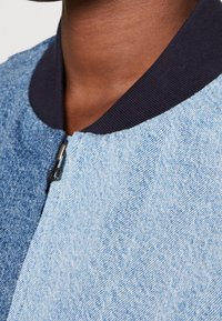 Dżinsowa kurtka bombka w jasnoniebieskim kolorze z teksturowanym wykończeniem, czarnym prążkowanym kołnierzem i widocznym zamkiem z przodu.