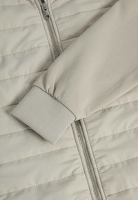 Giacca puffer beige chiaro con chiusura a zip. Presenta una texture trapuntata e polsini elastici a coste. Tessuto morbido con un finish setoso.