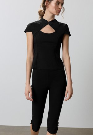 Une femme porte un haut noir à manches courtes avec une découpe au niveau de la poitrine et un pantalon noir court, sur un fond gris uni.