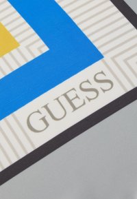 Design geometrico blu e giallo su tessuto con sfondo grigio; presenta la parola "GUESS" in lettere metallizzate dorate.