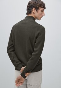 Dark green sweater with a high collar, textured ribbed design, and long sleeves. Model is viewed from the back, hands clasped.