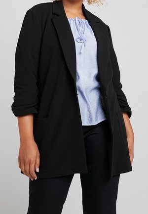 Personne portant un blazer noir ample avec des manches retroussées sur un chemisier bleu clair à col noué et un pantalon noir sur un fond uni.