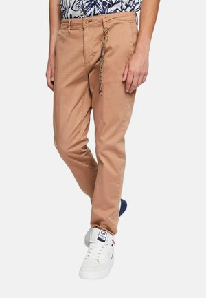 Pantaloni in cotone marrone chiaro con una vestibilità slim, dotati di un anello metallico con un dettaglio a catena decorativa. Abbinati a sneaker bianche.
