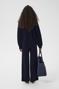 Άτομο με μακριά σγουρά μαλλιά που φοράει ναυτικό μπλε παντελόνι φαρδιού ποδός και πουλόβερ, κρατώντας μια μεγάλη τσάντα χειρός ναυτικού μπλε, στέκεται μπροστά σε λευκό φόντο.