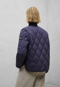 Marineblå quiltet puffer-jakke med diamantmønster, høj krave og et patch på venstre ærme, parret med lysebeige bukser.