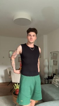 Top nero senza maniche con una texture liscia, abbinato a pantaloni corti verdi con tasche. Sullo sfondo si trova una sedia di colore chiaro e decorazioni.