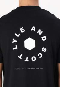 Μαύρο βαμβακερό t-shirt με μεγάλο λευκό γραφικό στο πίσω μέρος, περιλαμβάνοντας κείμενο και σχήμα εξαγώνου. Το κείμενο αναφέρει "LYLE AND SCOTT" και "SINCE 1874. FOOTBALL FOR ALL."