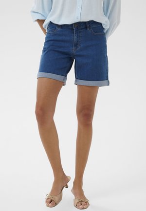 Kvinde iført opsmøgede mørkeblå denimshorts, lyseblå bluse og beige sandaler med hæl, stående med hænderne i lommerne.