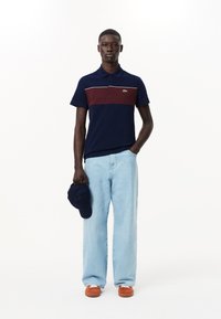 Laivastonsininen polo-paita, jossa on viininpunainen vaakaraita, lyhyet hihat ja kaulus. Vaaleansiniset leveät farkut, oranssit lenkkarit, pitää tummaa lakkia.