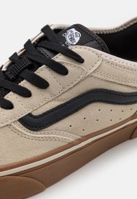 Baskets en suède beige avec bande en cuir noir, lacets noirs et semelle en caoutchouc marron. Présente des accents cousus et des détails perforés sur le côté.