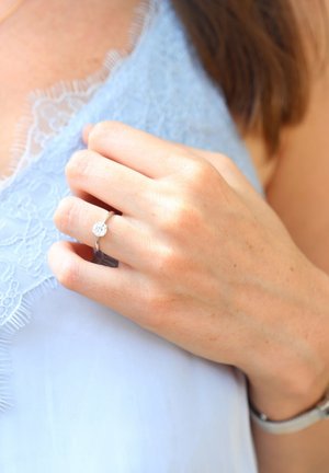 Bague en argent avec une gemme ronde et claire, portée sur une main tenant un tissu, avec des détails en dentelle bleu clair et une texture de peau lisse.