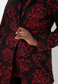 Černý blazer s vyvýšeným vzorem červených růží, s klopovým límcem a jednostranným uzavíráním na černý knoflík. Texturovaná tkanina zvýrazňuje vizuální zajímavost.