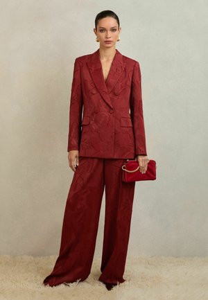 Vrouw draagt een getextureerde dieprode dubbelgerijpte blazer en wijde broek, houdt een rode clutch met gouden ketting vast, staand op een beige tapijt.