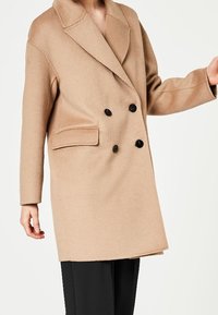 Mujer con un abrigo de lana beige de doble botonadura con botones negros y bolsillo con solapa, combinado con pantalones negros, frente a un fondo blanco.