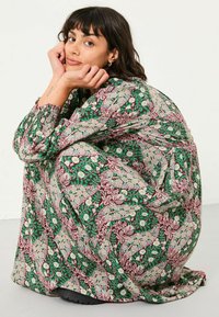 Langes, gemustertes Kleid mit grünen, burgunderfarbenen und cremfarbenen Blumenmustern, lockerer Schnitt, langen Ärmeln und elastischen Bündchen. Sichtbare schwarze Schuhe.