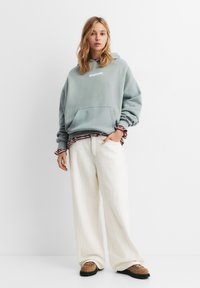 Miętowozielona, oversize'owa bluza z kapturem, z przednią kieszenią i białym napisem, nałożona na koszulę w paski. W połączeniu z białymi spodniami w szerokim kroju i brązowymi butami.