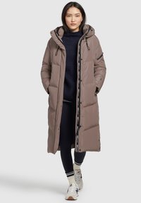 Lungo cappotto imbottito marrone con cappuccio, chiusura frontale con zip e tasche laterali. Presenta una texture morbida e un rivestimento interno con logo.