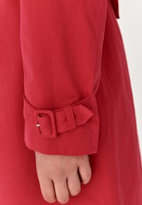Telaco rojo de abrigo con una textura suave, que presenta un puño con un detalle de cinturón. El puño está ligeramente vuelto hacia arriba, exponiendo la costura.