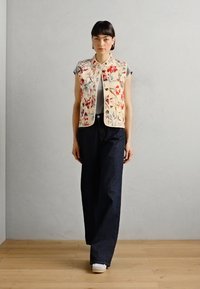 Blommönstrad broderad ärmlös väst med krämfärgad bakgrund, ihop med mörk denim vida byxor och vita sneakers på en trägolv.