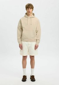 Felpa beige con coulisse, tasca anteriore, abbinata a pantaloni corti crema. Il modello indossa calze bianche e scarpe marroni scure, in piedi su uno sfondo neutro.