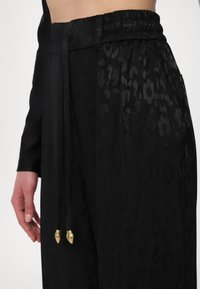 Zwarte broek met een subtiel luipaardpatroon, voorzien van een geplooide tailleband en een trekkoord met goudkleurige metalen uiteinden.