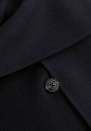 Materialul de lână de culoare bleumarin, cu o textură netedă, având un buton mare negru cu patru găuri și denumirea mărcii "Reiss" gravată pe el.