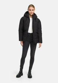 Young woman wearing a black hooded puffer jacket, white top, black skinny jeans, and black ankle boots standing against a white background.
