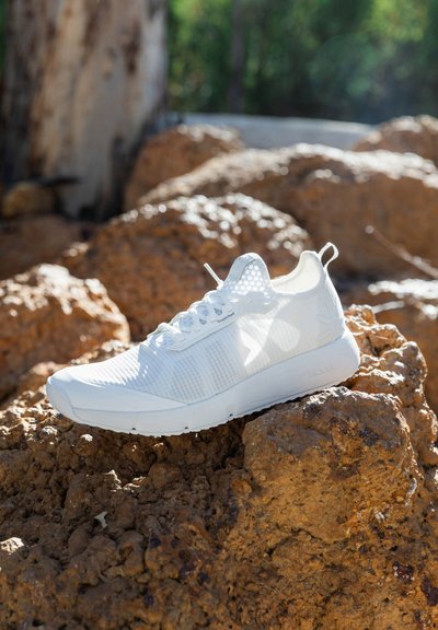 Chaussure de sport blanche avec une tige en maille respirante, surface texturée et semelle en caoutchouc flexible, posée sur un terrain rocheux.