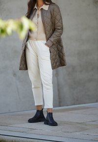 Cappotto lungo imbottito marrone con colletto crema, maglione beige a maglia, pantaloni bianchi a gamba affusolata e stivali Chelsea neri, in piedi su una superficie di pietra chiara.