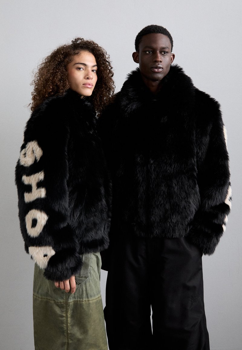 Dos personas llevan chaquetas de piel sintética negra; una de ellas tiene "SHOP" en color crema en la manga. Están combinadas con pantalones en verde oliva y negro.