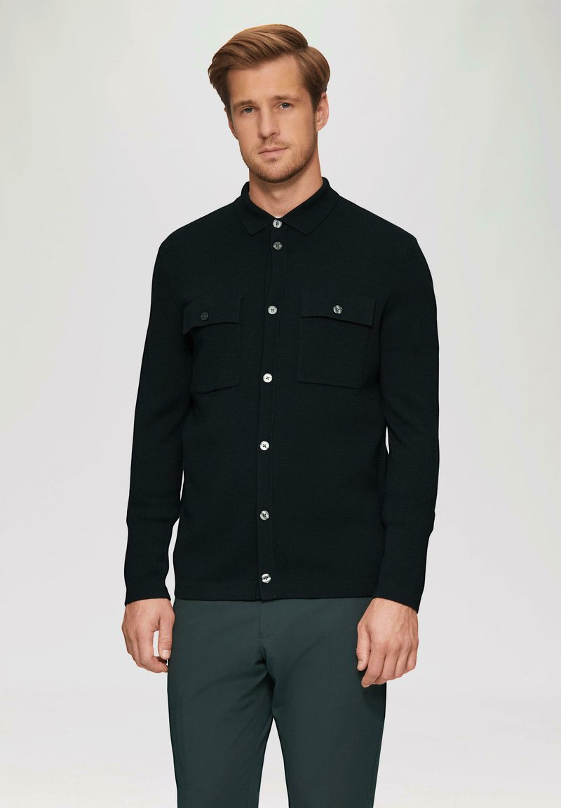 Zwarte lange mouwen shirt van een textuurstof, met een kraag, twee borstzakken en zilveren knopen langs de voorbies.