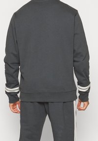 Sudadera gris con mangas largas, puños de canalé y rayas blancas en los codos. Diseño simple y textura suave, vista desde la parte de atrás.