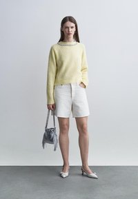 Nő áll, világos sárga pulóvert visel, melyen díszített nyakrészt visel, fehér rövidnadrágot, fehér magassarkút, és ezüst színű, bojtos kézitáskát tart.