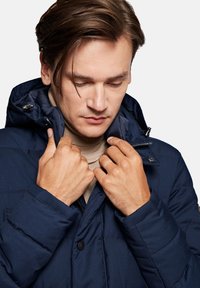 Granatowa pikowana kurtka z wysokim kołnierzem, zapięciem na guziki i kieszeniami na zamek. Teksturowany materiał oraz regulowany kaptur.