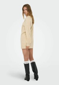 Gebreide, langemouwen jurk in lichtbeige. Kenmerken een ontspannen pasvorm, geribde manchetten en een ronde halslijn. Gemaakt in combinatie met kniehoge, zwarte leren laarzen.