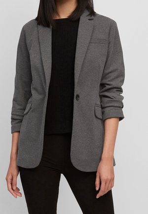 Nainen pukeutuneena harmaaseen bleiseriin, jonka hihat on kääräisty ylös, mustan ribbineuletakin ja mustien housujen kanssa, seisomassa yksinkertaisen harmaan taustan edessä.