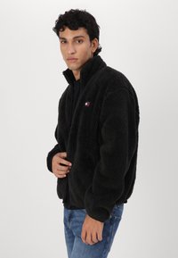 Chaqueta de forro polar negra y esponjosa con cuello alto y cierre de cremallera. Presenta un parche con el logo en el pecho. Combinada con vaqueros azules.