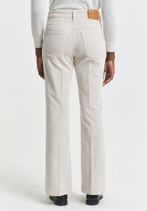 Person trägt hoch taillierte, cremefarbene Cordhosen mit Gesäßtaschen und einem hellbraunen Leder-Logoetikett, kombiniert mit schwarzen Schuhen und einem leichten, langärmligen Oberteil.